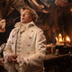 Pirate Stede Bonnet, wearing a fancy white outfit, is menaced by a dagger close to his eye.